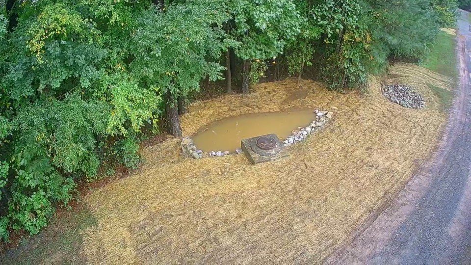 Newly constructed stormwater BMP with sediment basin and manhole structure surrounded by straw-covered soil and riprap, located at the edge of a wooded area.