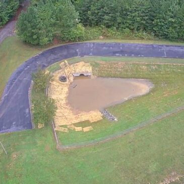 Aerial view of a newly constructed stormwater detention pond with outlet structure, riprap, straw erosion control, and surrounding paved access road.