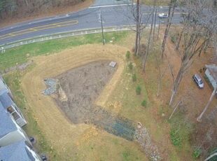 Aerial view of a stormwater drainage swale with riprap outlet leading toward a wooded residential area near a roadside.