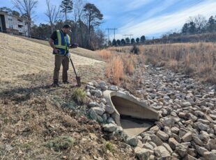 Stormwater professional inspecting a riprap-lined drainage outfall with a shovel and tablet, assessing erosion control near a culvert.