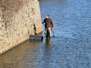 Stormwater technician standing in a retention pond inspecting an outlet pipe near a reinforced retaining wall.