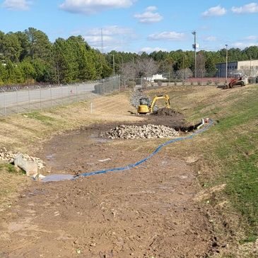 Stormwater detention basin under maintenance with exposed soil, riprap placement, and excavation equipment working on sediment removal and erosion control.